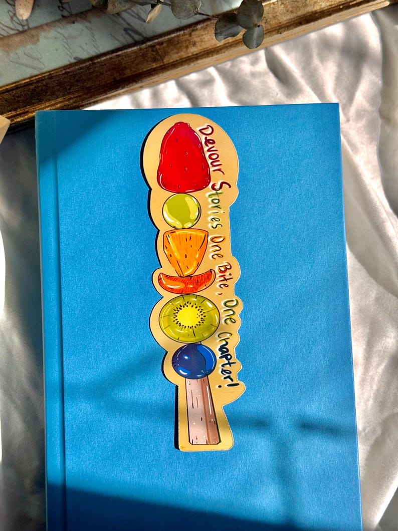 Pu&ograve; includere: Un segnalibro dorato con una pila di frutta colorata e il testo "Devour Stories One Bite, One Chapter!"