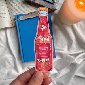 May include: A pink and red bookmark shaped like a bottle of strawberry soda. The bottle has a label that says "Strawberry Soda" and "Sip Read Repeat".