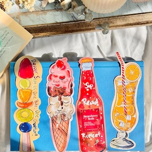 Pu&ograve; includere: Quattro segnalibri colorati con illustrazioni a tema estivo. I segnalibri raffigurano una bottiglia di soda alla fragola, un bicchiere di succo d'arancia, un ghiacciolo con frutta e un cono gelato. I segnalibri hanno il testo "Read Now's", "Sip Read Repeat", "Devour Stories One Bite, One Chapter!" e "Melt later!"