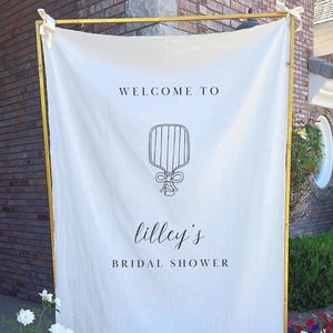 Può includere: Un banner di tessuto bianco con cornice dorata annuncia "Benvenuti all'addio al nubilato di Lilley". Il banner presenta un'illustrazione decorativa ed è legato con nastri bianchi. Lo sfondo comprende un muro di mattoni e vegetazione.
