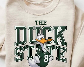 Puede incluir: Una sudadera beige con un gráfico verde y blanco de un pato de dibujos animados que lleva una camiseta con el número 8. El texto "The Duck State" está impreso en letras verdes sobre el pato.