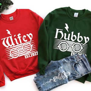 May include: A red sweatshirt with the word "Wifey" in white, stylized with glasses and the text "EST.2022". A green sweatshirt with "Hubby" in white, glasses, and "EST.2022". Blue jeans are folded beneath.