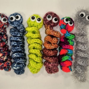 May include: Six crocheted worm toys with googly eyes. The worms are in various colors, including red, blue, green, brown, and gray.