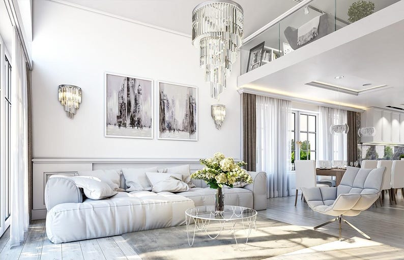 May include: A living room featuring a large white sectional sofa, a glass coffee table with a flower vase, a tufted gray armchair, and a tiered crystal chandelier. Two framed abstract paintings hang on the wall above the sofa.