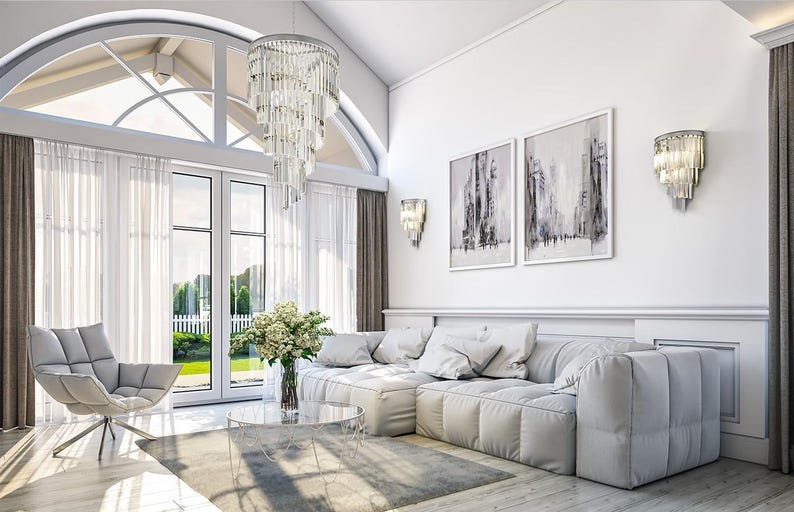 May include: A living room featuring a large white sectional sofa, a gray armchair, a round glass coffee table with a flower vase, a tiered crystal chandelier, two wall sconces, and two framed abstract paintings on a white wall.