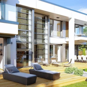 May include: Exterior view of a modern two-story house with large glass windows and doors. Two dark gray chaise lounges with light blue cushions sit on a wooden deck. A dining table and chairs are visible on a covered patio.