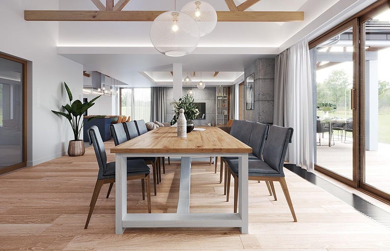 May include: A long rectangular dining table with a light wood grain top and a white base, surrounded by eight dark gray upholstered chairs with wooden legs. The table is positioned in a bright, open-plan room with light wood flooring.