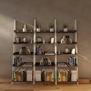 K&ouml;nnte beinhalten: Ein dreiteiliges Leiterregal mit wei&szlig;en, schr&auml;gen Seitenst&uuml;tzen und dunkelbraunen horizontalen Regalb&ouml;den. Die Regale sind mit verschiedenen B&uuml;chern, kleinen gr&uuml;nen Topfpflanzen, wei&szlig;en Tierfiguren und gerahmten Bildern gef&uuml;llt.