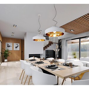 Pu&ograve; includere: Una sala da pranzo con un lungo tavolo in legno e sedie bianche, due lampade a sospensione bianche con interno dorato e una grande finestra. Lo spazio presenta pavimenti in legno chiaro, accenti neri e una scala sullo sfondo.
