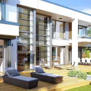 May include: Exterior view of a modern two-story house with large glass windows and doors. Two dark gray chaise lounges with light blue cushions sit on a wooden deck. A dining table and chairs are visible on a covered patio.