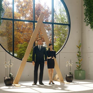 May include: A person in a black suit with a white shirt and striped tie stands next to a person in a black dress. They are positioned in front of a large circular window with a wooden A-frame structure, flanked by two potted orchids and a corn plant.