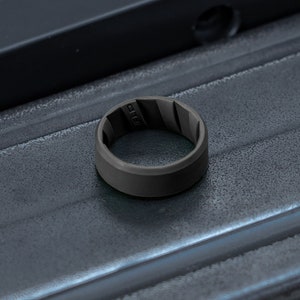 Pode incluir: Um anel de silicone cinza-escuro fosco com um design interior texturizado e ranhurado. A banda tem uma superf&iacute;cie exterior lisa e s&oacute;lida e est&aacute; posicionada sobre um objeto retangular met&aacute;lico escuro com um acabamento industrial ranhurado.