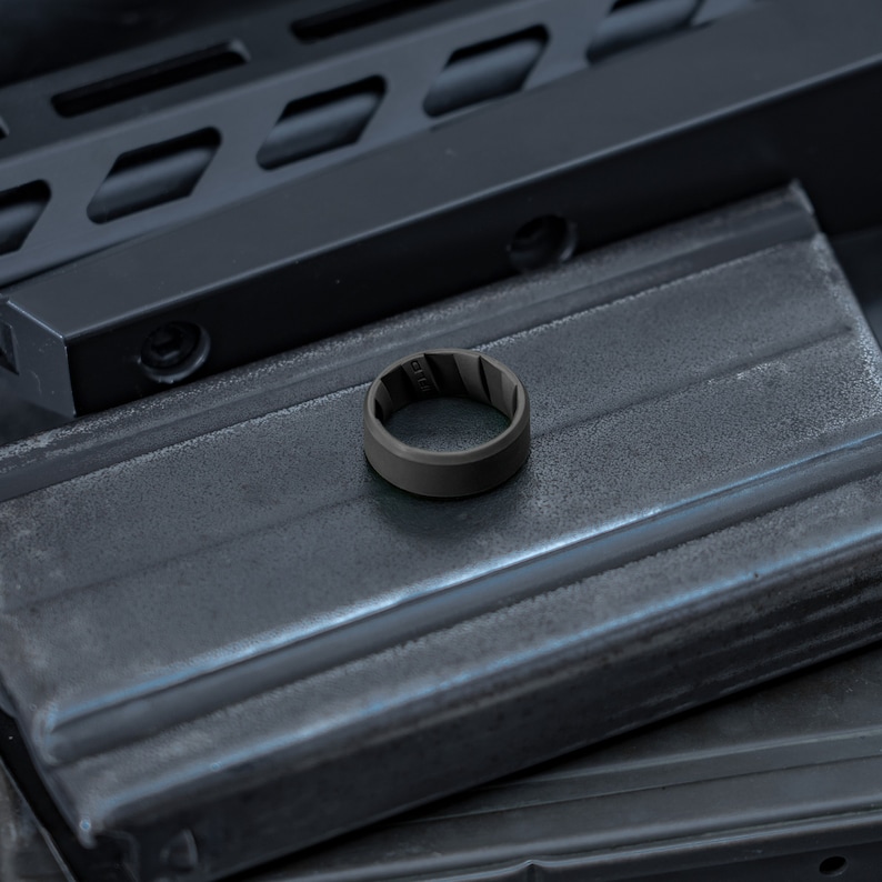 Pode incluir: Um anel de silicone cinza-escuro fosco com um design interior texturizado e ranhurado. A banda tem uma superf&iacute;cie exterior lisa e s&oacute;lida e est&aacute; posicionada sobre um objeto retangular met&aacute;lico escuro com um acabamento industrial ranhurado.