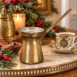 May include: A brass coffee pot with a long handle and a wide, flared rim. The metal surface shows a weathered, aged patina. The pot is positioned on a marble tray alongside a white porcelain teacup with a green and gold floral pattern.