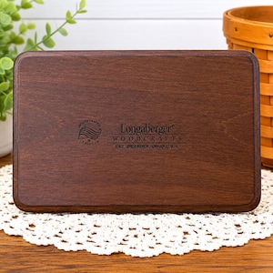 May include: A rectangular dark brown wooden lid with rounded corners. The center features a black stamped logo that reads Longaberger WOODCRAFTS EST. DRESDEN, OHIO U.S.A. along with a circular icon containing the text PROUDLY CRAFTED IN THE U.S.A.