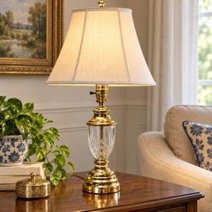May include: A table lamp featuring a polished gold-tone metal base with a clear, cut-glass center section and a flared, off-white fabric lampshade. The lamp has a classic silhouette with a tiered base and a small, round finial at the top.