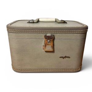 May include: A vintage gray vanity case with a cream-colored handle and a gold-toned clasp. The case has a rectangular shape with rounded corners and decorative stitching. The brand name is visible on the front.