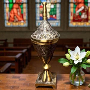 May include: A tall, metallic gold-toned incense burner featuring a textured, conical lid topped with a slender finial. The body has a tapered, ribbed design and a square base. Hebrew characters are engraved along the rim of the lid.