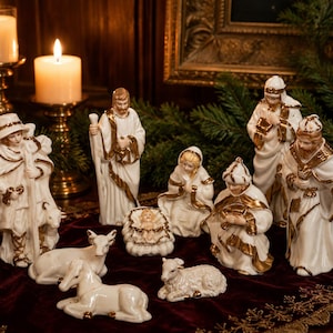 May include: A porcelain nativity set featuring white figurines with gold trim, including a central infant in a manger, figures of adults, a shepherd, and animals such as a sheep and a donkey, all arranged on a dark red velvet fabric surface.