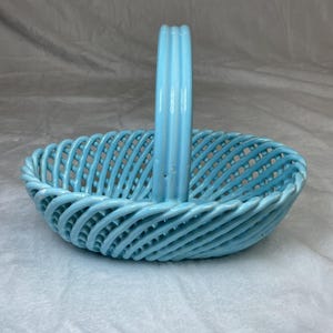 May include: A light blue ceramic basket with a woven design and a vertical handle. The basket is oval-shaped and features a glossy finish. The handle is ribbed and adds to the basket's decorative appeal.