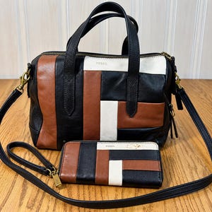 May include: A Fossil handbag and matching wallet featuring a patchwork design of black, brown, and white leather panels. The handbag has two top handles, a detachable shoulder strap, and a zipper closure. The wallet includes a wrist strap.