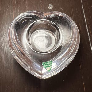 May include: Clear glass heart-shaped candle holder with a circular indentation in the center. The candle holder has a label that reads "Orrefors Sweden". The glass is transparent, allowing light to pass through.