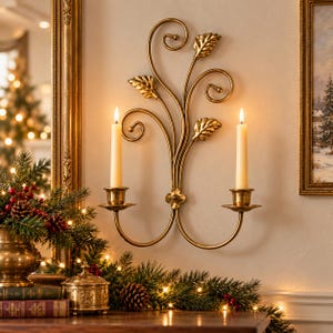 May include: A gold-toned metal wall sconce featuring two candle holders, decorative swirling metal vines, and three leaf accents. Two cream-colored taper candles are inserted into the cups, with a central flower-shaped metal detail.