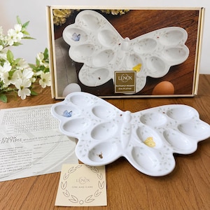 May include: A white ceramic egg platter shaped like a butterfly with twelve oval indentations. The platter features delicate gray leaf patterns, a blue butterfly, a yellow butterfly, and a bee. The box is labeled LENOX BUTTERFLY MEADOW EGG PLATTER.
