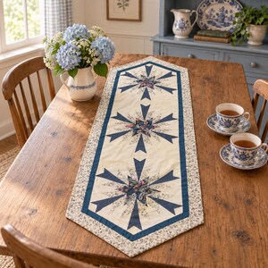 May include: A hexagonal table runner featuring three central star motifs in navy blue and floral fabric on a cream background, bordered by a small-scale floral print. The runner is placed on a wooden table with a blue and white ceramic pitcher.