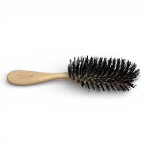 May include: A hair brush with a light beige handle and black bristles. The brush has a curved handle and a rectangular bristle head. The handle has text printed on it. The brush is designed for grooming and styling hair.