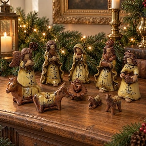 May include: A ceramic nativity set featuring figures in yellow robes with dark brown floral patterns, including an angel, three figures, a baby in a manger, two donkeys, and two sheep, all arranged on a wooden surface with festive greenery.