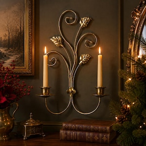 May include: A gold-toned metal wall sconce featuring a symmetrical design of swirling vines and three stylized leaves. The fixture includes two circular candle cups, each holding a tall, slender, cream-colored taper candle with a lit flame.