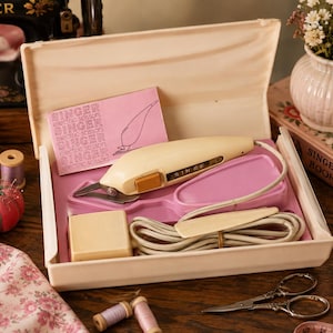 May include: A vintage Singer electric scissors set in a cream-colored plastic case. The set includes a cream-colored electric cutter with an orange switch, a power cord, and a pink instruction card with the text SINGER electric scissors.