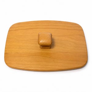 May include: A rectangular wooden lid with rounded corners and a smooth, light brown finish. The lid features a centered, square-shaped wooden knob handle with a slightly tapered base, providing a simple grip for lifting the cover.
