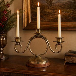May include: A brass candelabra featuring a central circular frame and three candle holders, each containing a lit white taper candle. The metal structure rests on a circular wooden base, showcasing a polished finish with curved arms.