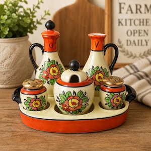 May include: A ceramic condiment set featuring an orange and cream tray holding two pitchers labeled O and V, a lidded sugar bowl, and two shakers. Each piece is decorated with hand-painted red, yellow, and green floral motifs and black accents.