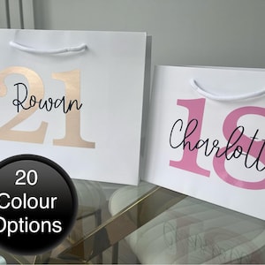 May include: Two white gift bags with handles. The bag on the left has the number 21 in gold foil with the name Rowan written in black script. The bag on the right has the number 18 in pink foil with the name Charlotte written in black script.  There are 20 colour options available.