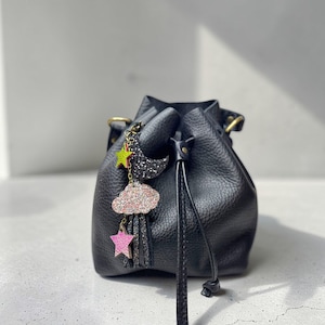 May include: Black leather drawstring bag with a decorative charm featuring a crescent moon, cloud, and star. The charm has glitter and pink accents. The bag has a textured surface and a drawstring closure.