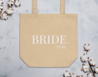 May include: A beige tote bag with white handles and the text "BRIDE TO BE" printed on the front.