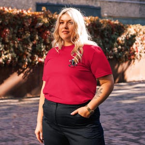 May include: A red t-shirt with a small graphic on the chest. The graphic appears to be a cartoon character. The shirt is made of a soft material and has short sleeves. The person is wearing black pants and a gold watch.