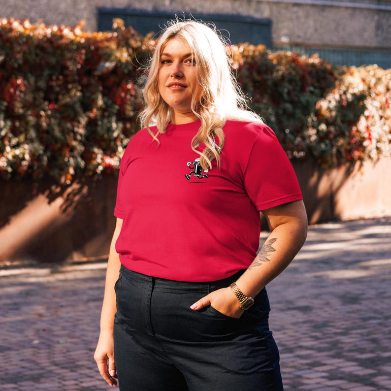 May include: A red t-shirt with a small graphic on the chest. The graphic appears to be a cartoon character. The shirt is made of a soft material and has short sleeves. The person is wearing black pants and a gold watch.