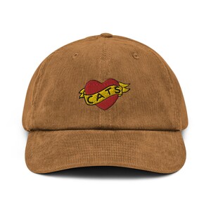 Puede incluir: Una gorra de béisbol de pana marrón con un corazón rojo bordado y la palabra "CATS" en amarillo.
