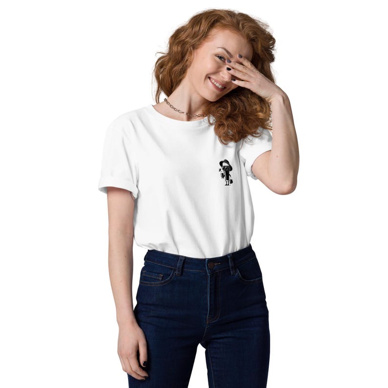 Puede incluir: Camiseta blanca con un peque&ntilde;o gr&aacute;fico negro en el pecho. El gr&aacute;fico es una figura estilizada. La camiseta se combina con vaqueros azul oscuro. La modelo sonr&iacute;e y se cubre la cara con la mano.