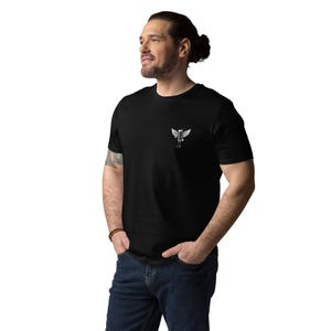 May include: Black t-shirt with a small white graphic of a winged figure on the left chest. The t-shirt is made of a soft material and is being worn by a person in blue jeans.