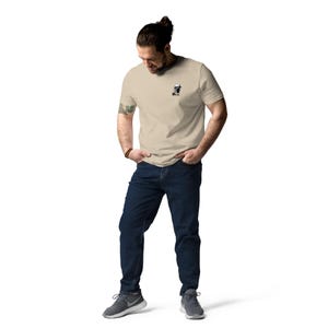 Puede incluir: Camiseta beige con un peque&ntilde;o gr&aacute;fico negro en el pecho izquierdo. Se combina con vaqueros azul oscuro y zapatillas deportivas grises. La persona est&aacute; de pie con las manos en los bolsillos.