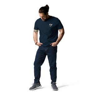 May include: A navy blue t-shirt with a small graphic on the chest. The person is wearing dark wash jeans and gray athletic shoes. The person is standing with their hands in their pockets.