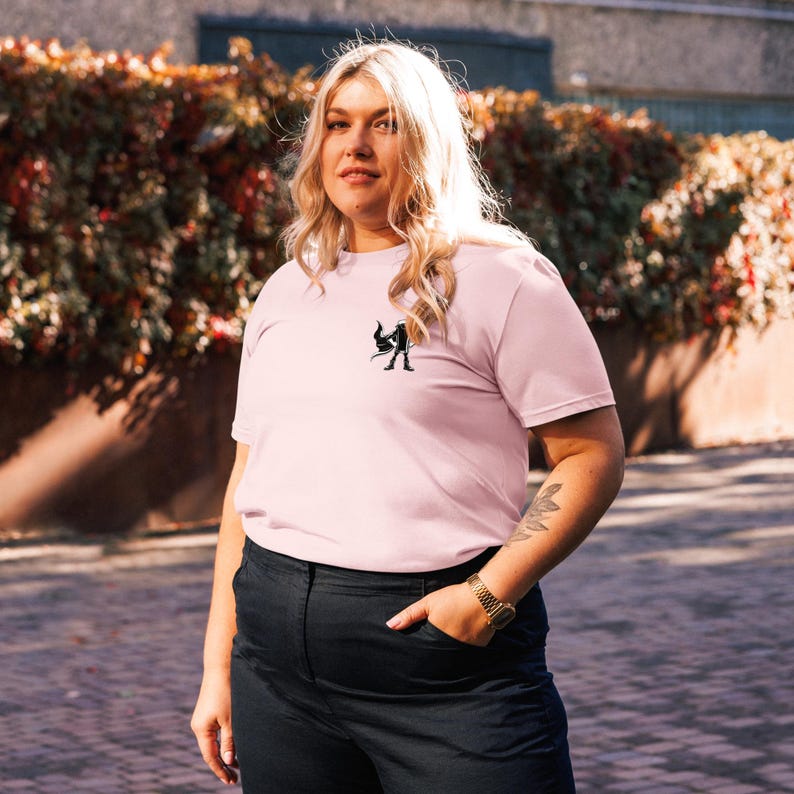May include: A pale pink t-shirt with a small black graphic on the chest. The shirt has short sleeves and a crew neck. The person is wearing black trousers and a gold watch. The background features green and brown foliage.