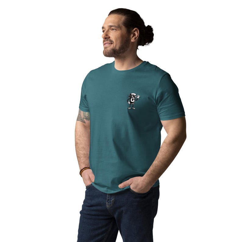 Puede incluir: Camiseta de color turquesa con un peque&ntilde;o personaje de dibujos animados que lleva un sombrero blanco y la letra "G". El personaje est&aacute; saludando. La camiseta est&aacute; hecha de un material suave y la lleva una persona.