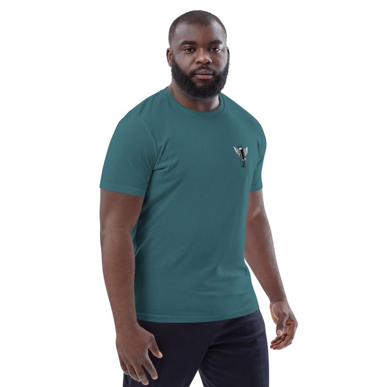 May include: Teal short-sleeve t-shirt with a small, embroidered design on the upper left chest. The design appears to be a stylized bird or wing shape in black and white. The shirt is made of a soft material.