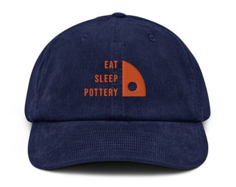 Może przedstawiać: Czapka bejsbolowa z niebieskiego, prążkowanego materiału typu corduroy z haftowanym pomarańczowym logo z napisem "Eat Sleep Pottery" i stylizowaną pomarańczową kołem garncarskim.